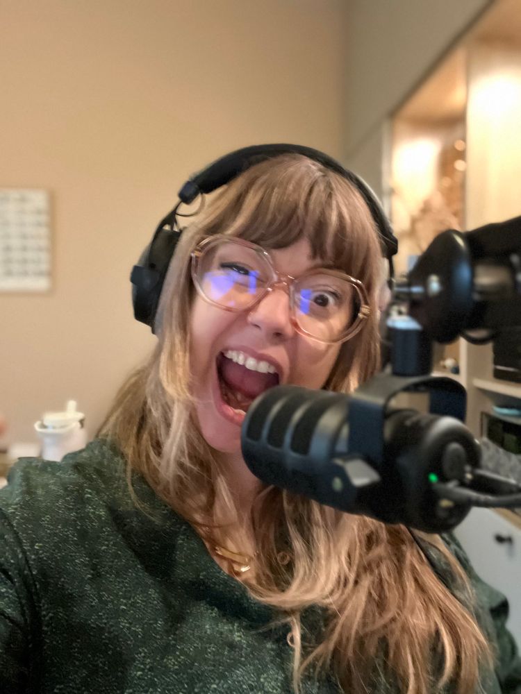 A white middle aged blonde woman with bangs and glasses pulls a screaming face, with one eye closed. She’s wearing headphones with a mic in front of her. Oh and a green hand made blouse.  