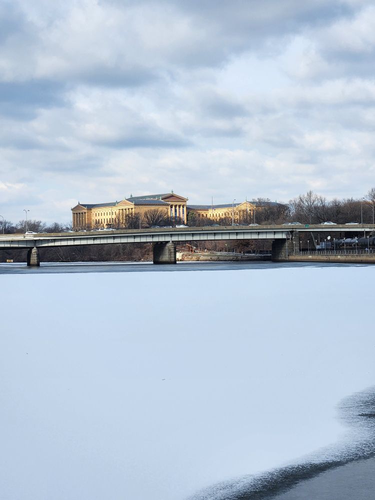 Photo of a building in Greek classical style, under clouds, lit by sunshine from the left. In the foreground is a snow-covered river. A highway bridge runs just below the building in this view.