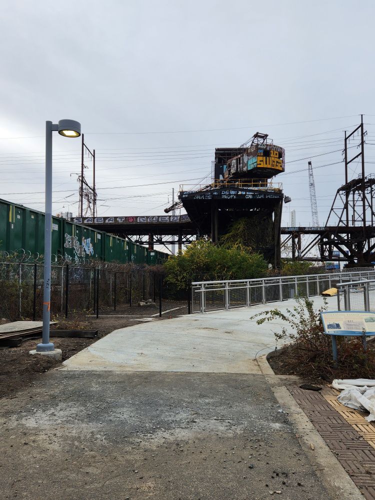 Photo of a concrete walkway between a macadam surface and an elevated walkway with side railings, plus a railroad bridge and graffiti tagged elevated crane.