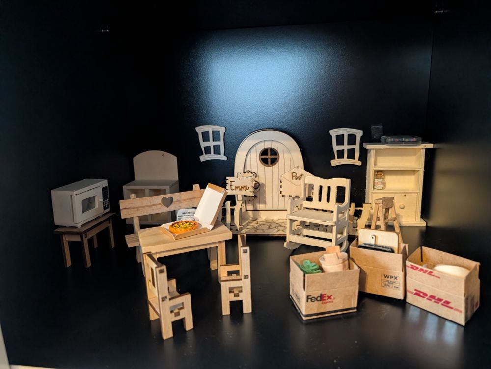 Ein Regalfach mit kleinen Holzmöbeln, einer Tür und drei Umzugskartons. Links ist ein Essbereich eingerichtet mit Bank, Tisch und zwei Stühlen. Dahinter eine Mikrowelle. Auf dem Tisch steht ein Karton mit Pizza drin. Rechts daneben stehen die Kartons, dahinter ein Schaukelstuhl, eine Trittleiter und ein Regal. Vor der Tür im Hintergrund ist ein Briefkasten und ein Namensschild mit dem Namen "Emmi". 