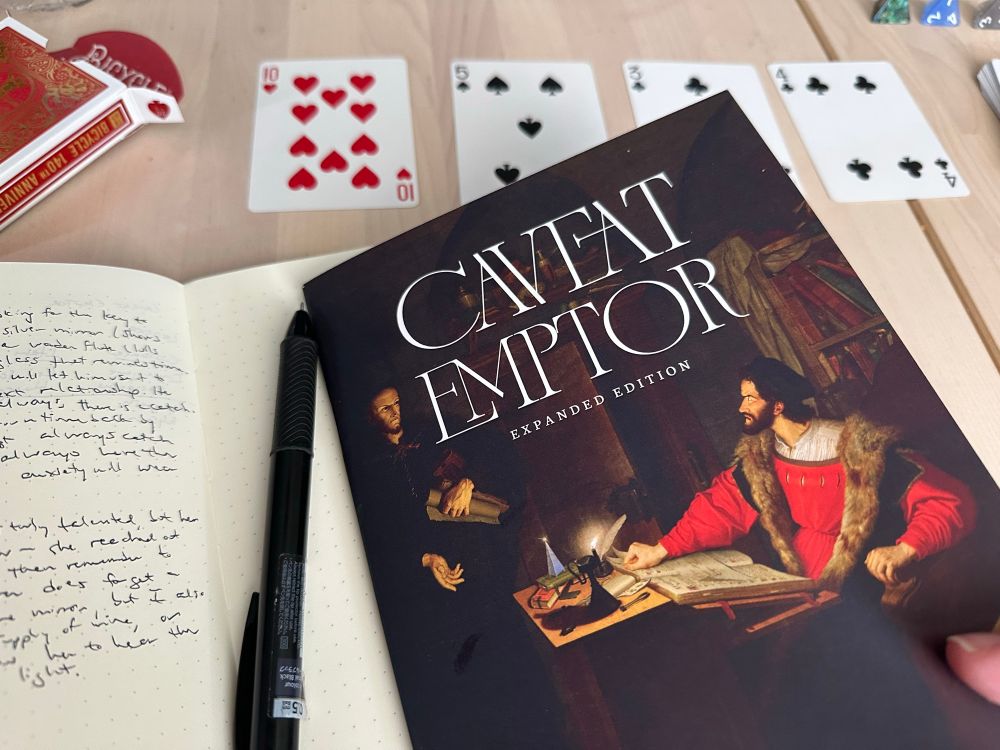 Caveat Emptor book on top of a journal with writing in it, with playing cards arranged in the background. 