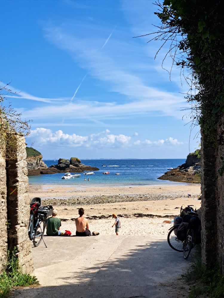 Vue sur une plage de Belle-île-en-mer, entre deus murs, la plage de sable s'étend, puis des rochers de part et d'autre, puis la mer, puis le ciel bleu. Deux personnes sont assises sur le sables et regardent l'horizon, un enfant joue, la plage sauvage est presque déserte. 