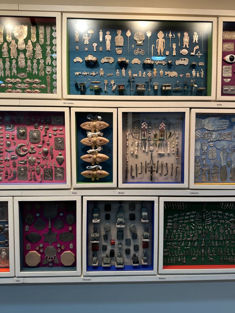 trinkets, jewelry, and other little heirlooms from Sri Lanka, Italy, Brazil, Lebanon, Iran, Ethiopia, and Mexico, arranged in frames for each country. some are carved out of bone, and some are pressed into silver