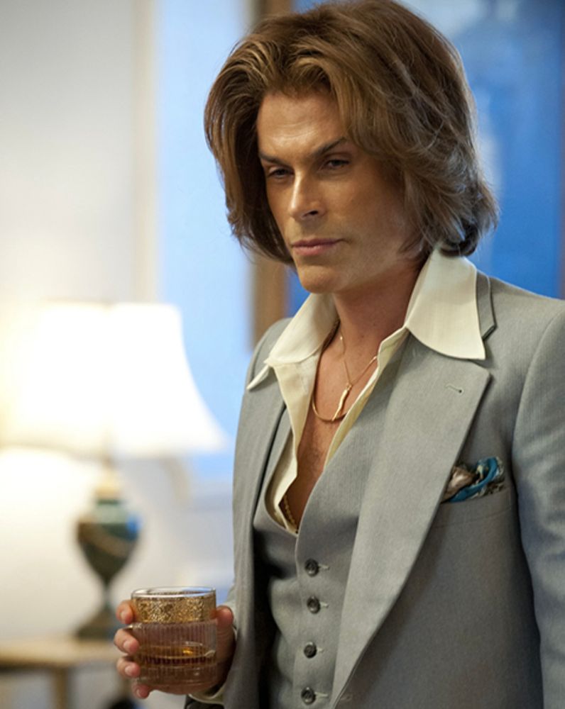 Rob Lowe in the film Behind The Candelabra with makeup effects applied to his face to make him look like he’s had lots of plastic surgery. He’s wearing a light grey suit and is holding a drink. 
