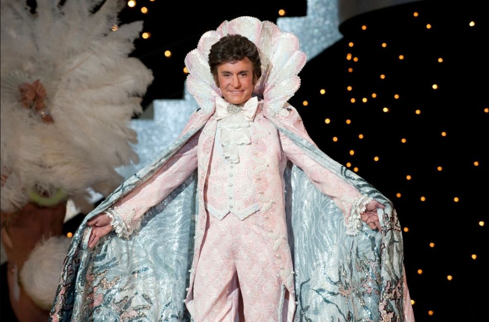 Michael Douglas as Liberace wearing a light pink suit with a cowl and blue cape. 