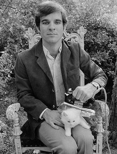 A black and white photo of comedian Steve Martin in the 1970s ironing a white kitten sitting outside in front of bushes. 
