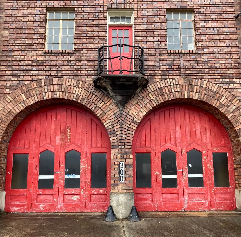 Front view of a brick fire station building built in 1927. Two tall wood doors with circular tops are painted red. These doors are wide, designed to accommodate fire trucks. Above these doors is a small balcony.