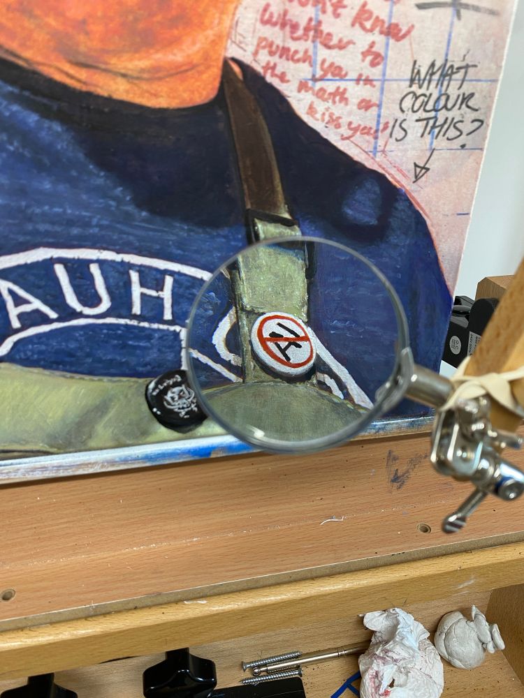 A detail of the previous picture shows a magnifying glass attached to a Mahl stick which shows a close up of a no to AI badge attached to my painting apron.
