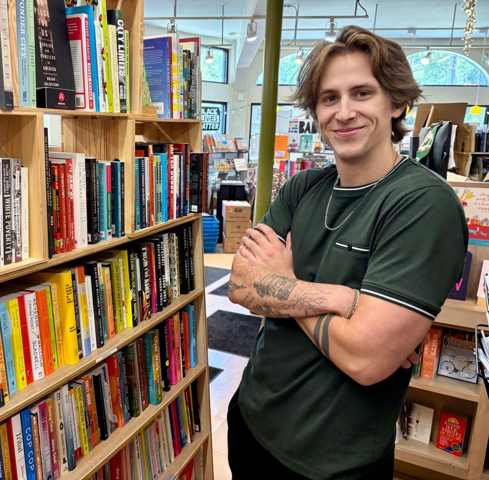 Micah in a green shirt standing in front of a book case with his arms crossed, tattoos on arms, a chain necklace, and short-ish hair parted in the middle.