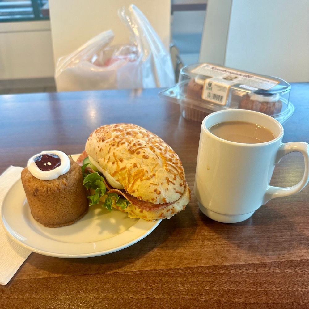 Kuvassa lautasella runebergintorttu ja sämpylä, vieressä kuppi kahvia, taustalla 2 kpl rasia runebergin torttuja ja muovikassillinen pullaa