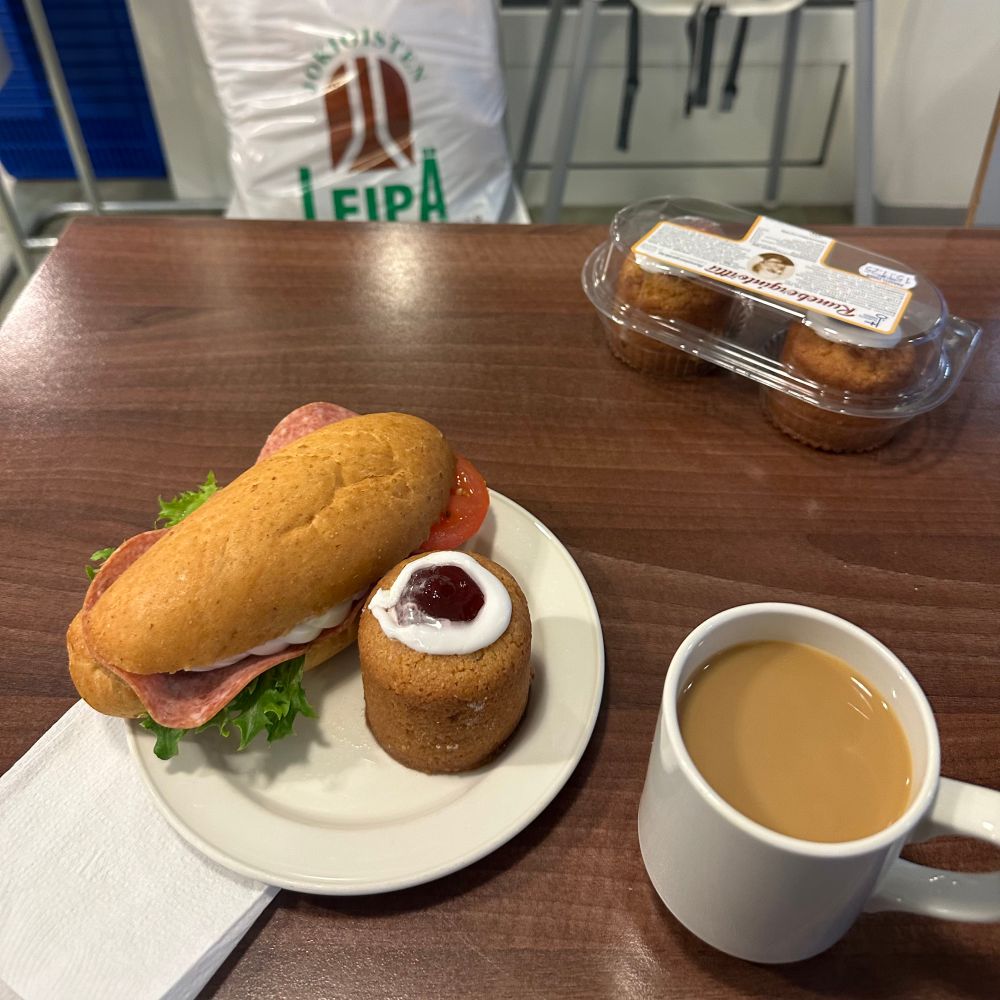 Sämpylä, runebergintorttu, kuppi kahvia, tortturasia ja vastapäisellä tuolilla pullakassi, jonka kyljessä lukee jokioisten leipä