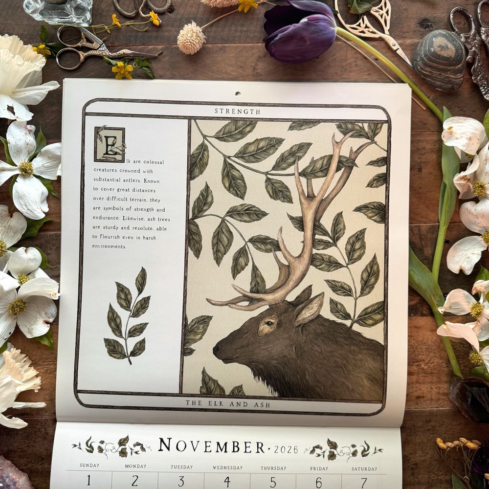 Photo of the 2026 Woodland Wardens Calendar, on a wooden desk surrounded by flowers. The calendar is open to November, which shows an elk and leaves. 