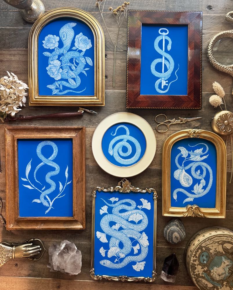 Photo of six blue and white paintings of snakes on a wooden table. A jumble of different frames, some rectangles, some rounded top rectangles, some ornate, one circular. 
