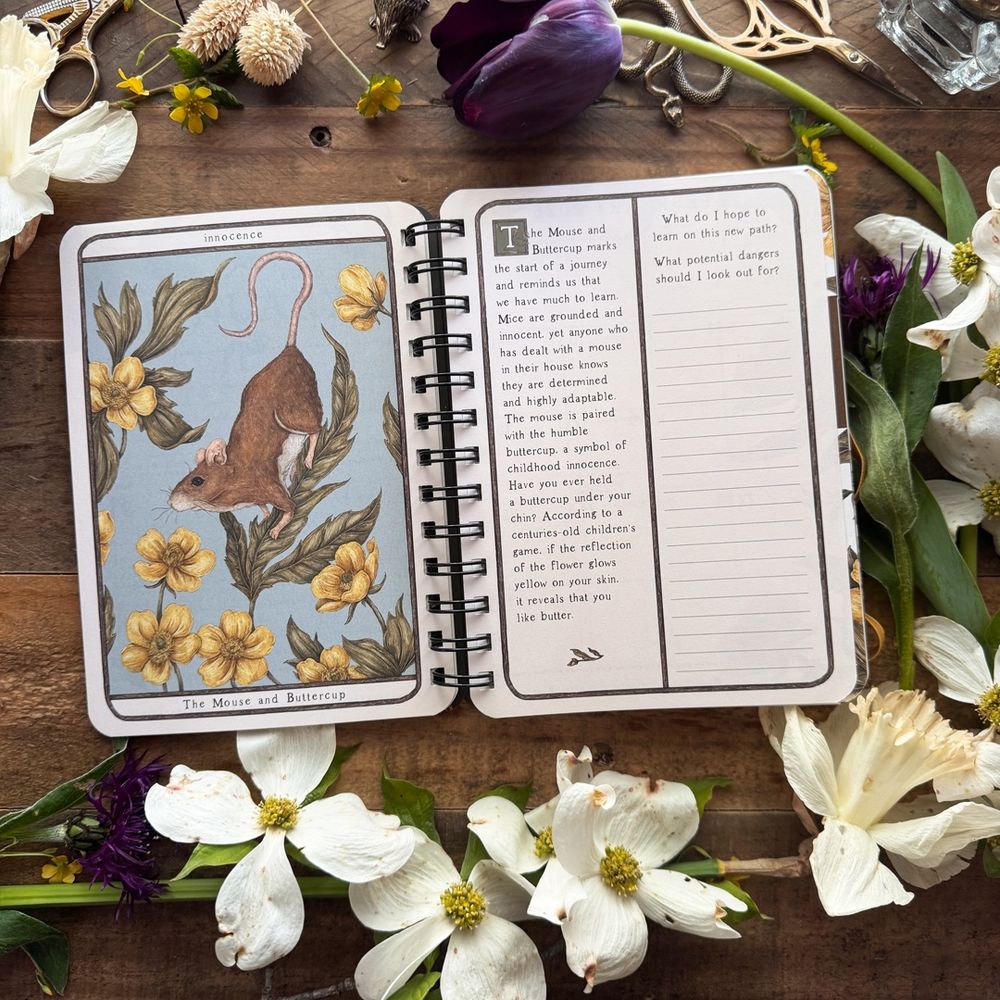 Photo of the 2026 Woodland Wardens Weekly Planner, on a wooden desk surrounded by flowers. The planner is open to a page showing an illustration of a mouse with buttercup flowers.  
