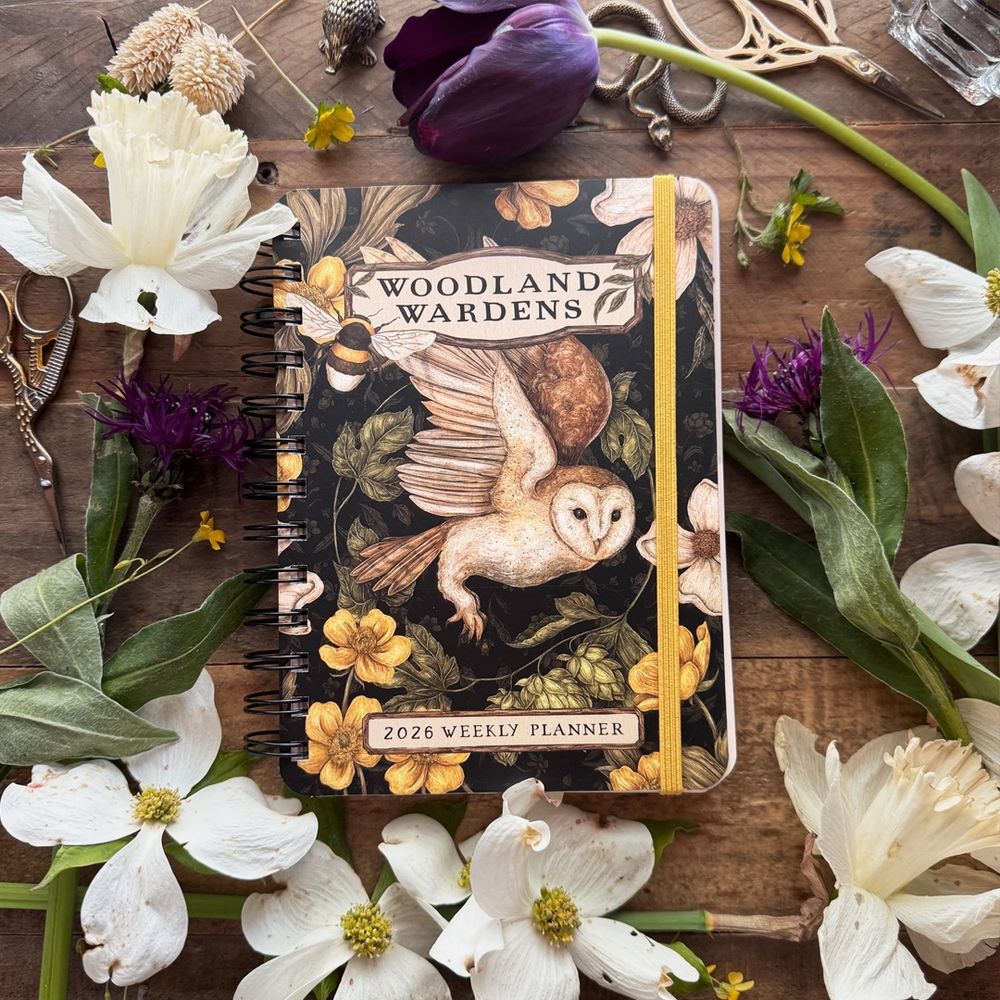 Photo of the 2026 Woodland Wardens Weekly Planner, on a wooden desk surrounded by flowers 