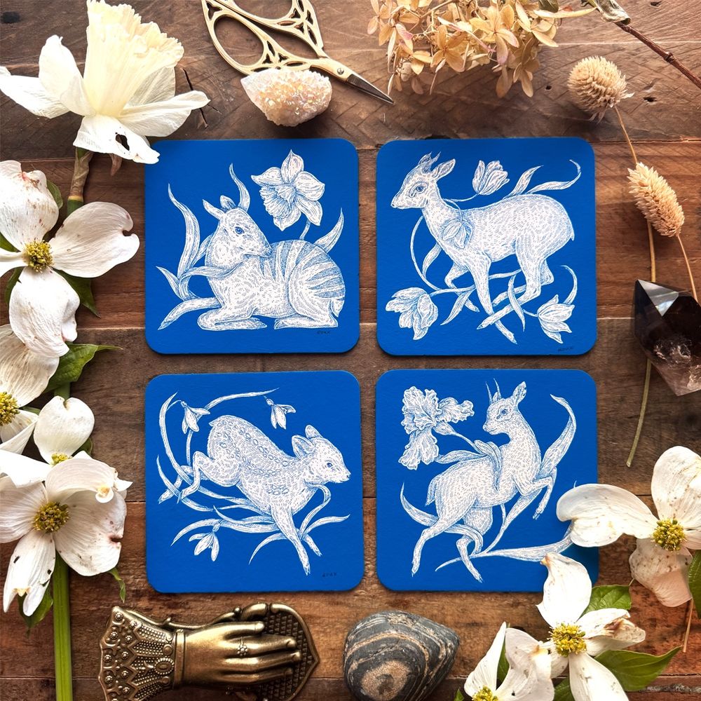 Photo of four small square paintings arranged on a wooden table surrounded by flowers. The background of each piece is blue, and they show a blue and white drawing of a zebra duiker with a daffodil, a dik-dik with tulips, a pudu with snowdrops, and a klipspringer with an iris. 
