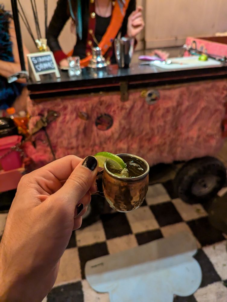 A photo of my left hand holding a miniature Moscow mule, complete with a wedge of key lime to go with the tiny (probably 1/6th size) mule cup. 