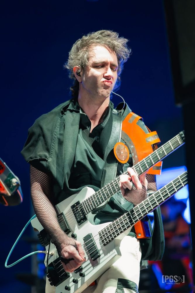 A photo of myself playing a white double-neck guitar/bass guitar wearing a black cloak, orange pauldron, white and black cargo pants, and fishnet shirt. My hair is short and I don't have a beard, and it's very windy so my hair is all pushed to one side.