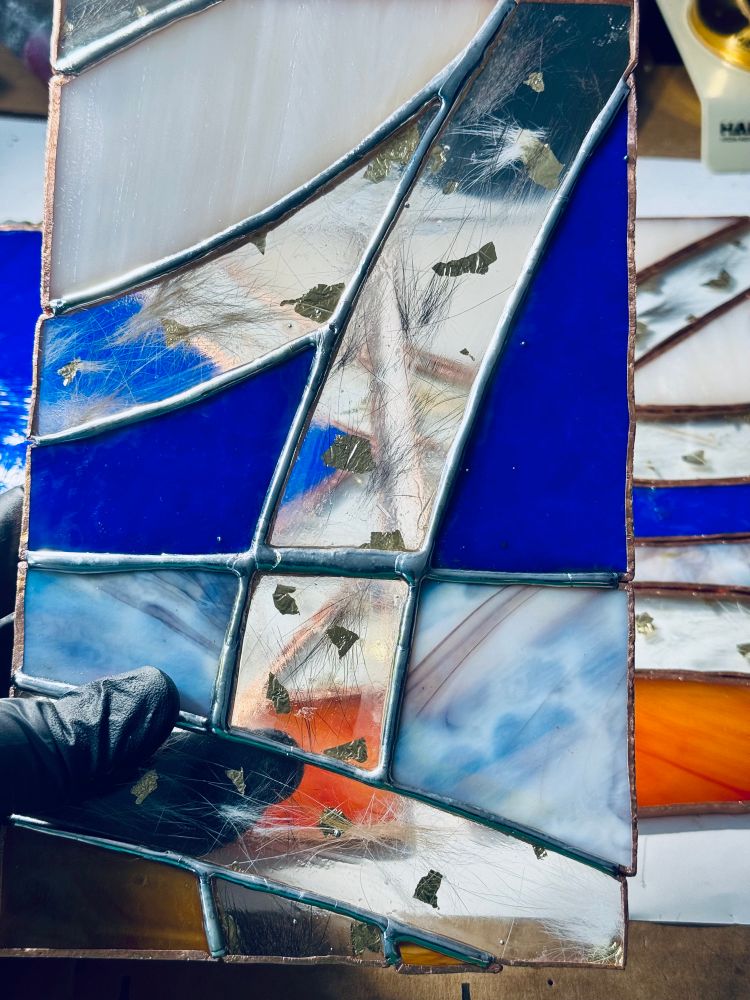 One of the side panels soldered together. There stripes of clear glass with cat fur and gold leaf pressed between them. The other glass colors are a peachy white, blue, grey stipple and wispy orange. You can see parts of the other panels unsoldered in the background 