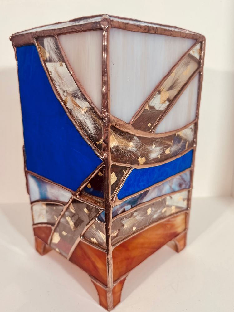 A view of the back and side of the vertical glass container. You can see a variety of glass. Orange, blue, grey and white. There are pieces of clear glass that have fur and gold leaf pressed in them.