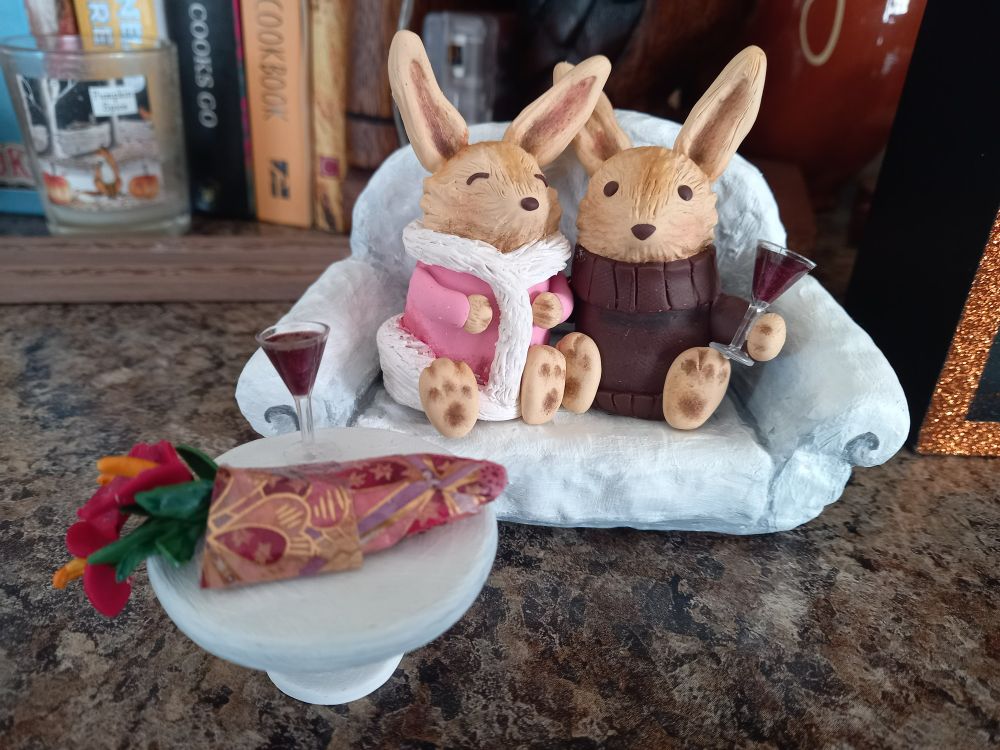 A sculpture of two bunnies cuddling on a sofa, they are wearing chunky jumpers and have a glass of wine each. The artist is Manen Lyset who also writes amazing stories.