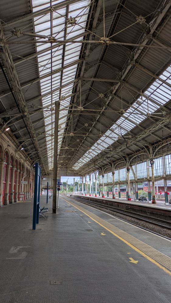The image shows a view of a railway station, looking down the platform to the tracks beyond. The platform is marked with accessibility strips (yellow and bumpy) and the station roof is old and Victorian, vaulted and beamed in cast iron and a haven for pigeons. 