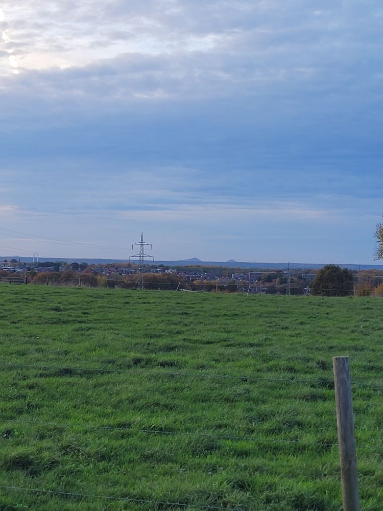 A green field and beyond that is an endless seeming expanse of very flat land with two small round hills far off in the distance. They look like islands in an ocean