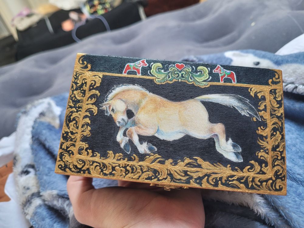 Top of a wooden box featuring a leaping fjord horse on colored pencil and a painted border with a floral and dala horse theme