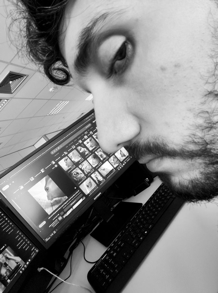 Selfie em preto e branco do meu rosto barbado em um ângulo pegando de perfil mas um pouco por trás da cabeça, estou olhando de canto de olho em direção à câmera, tenho olheiras; minha franja cacheada aparece meio bagunçada e com um "pega rapaz" se destacando dela; ao fundo, um monitor com uma página do Google Imagens aberta com fotos de memes de cavalos na mesma posição que copiei na selfie.