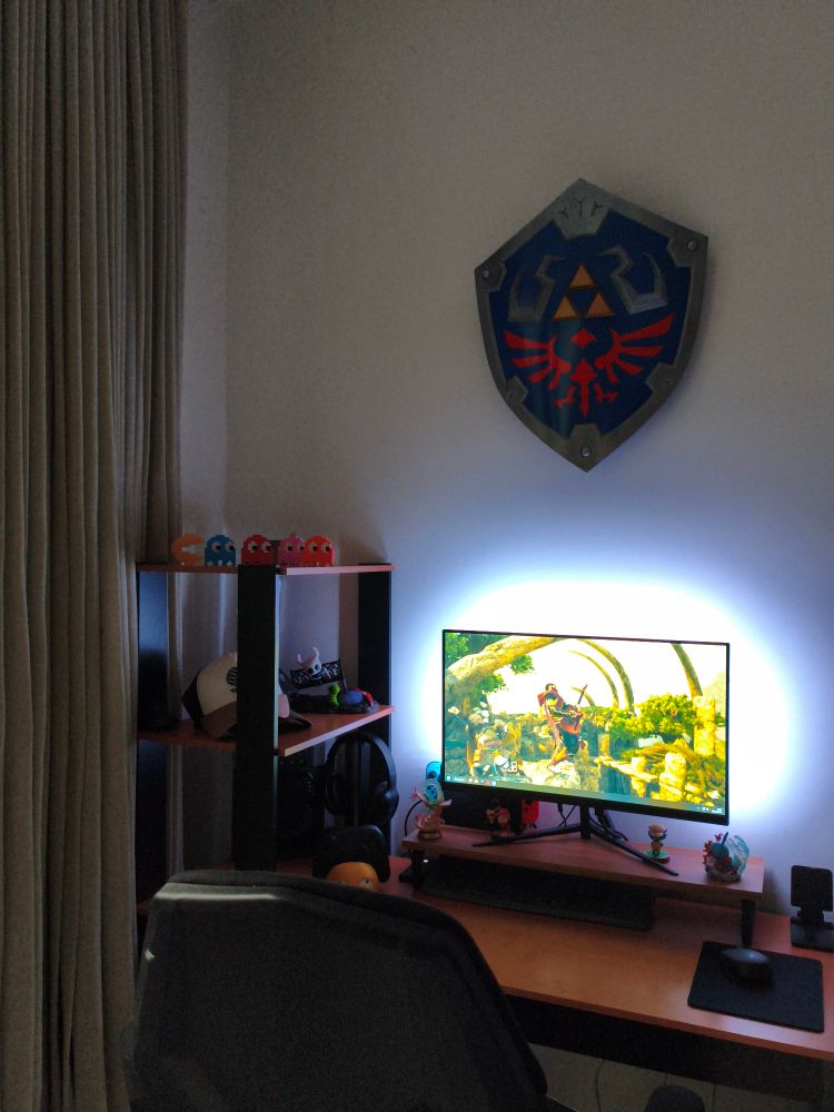 Foto de uma escrivaninha de madeira com prateleiras. Nas prateleiras tem decorações de personagens de Pacman, Hollow Knight e Pokémon, e bonés. Na parte maior da escrivaninha está o monitor do computador, um Nintendo Switch atrás dele e uma luz ligada ao fundo. Ao lado esquedo da foto aparece a cortina. Em cima de tudo está um escudo Hylian preso na parede.