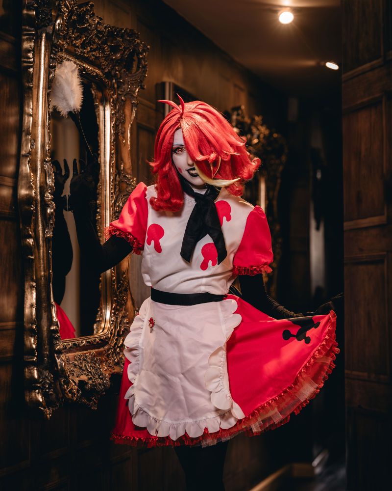 girl in niffty from hazbin hotel cosplay, next to mirror holding a feather duster, looking at the camera 