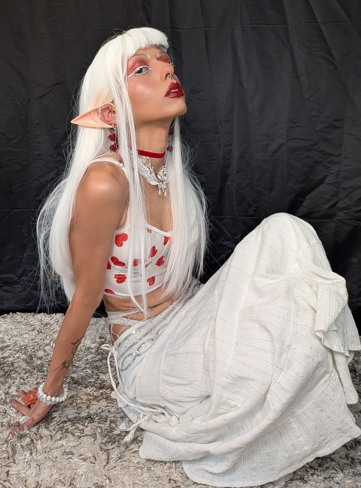 White haired elf girl with red lips white eyeliner and mascara, red eyeshadow. Wearing a white top with red hearts, and a white skirt 