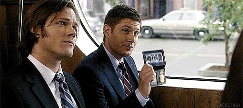 Dean Winchester (Jensen Ackles) from Supernatural. He is seated at a diner, flashing his (fake) FBI badge. With his dark navy suit he is wearing a tie that features diagonal stripes of crimson, navy, and silver/blue.