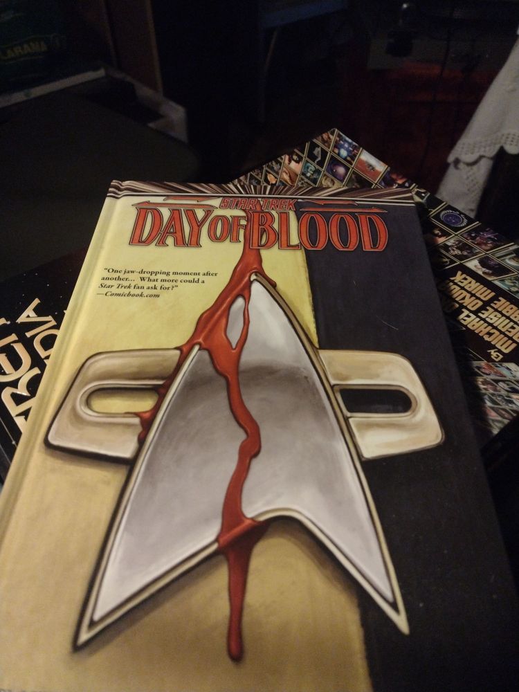 A hard-bound graphic novel with a vertically striped ¾ yellow & ¼ black front cover. On the top is written Star Trek. The line below it in larger writing states Day of Blood in all caps. Flowing down from the top center is a trail of blood which drips all the way down the front of a Voyager-style comm badge and back onto the yellow background. A tiny bit of blood also can be seen on the side of the badge. On the left hand side is a quote from comicbook.com - "One jaw-dropping moment after another... What more could a Star Trek fan ask for?'