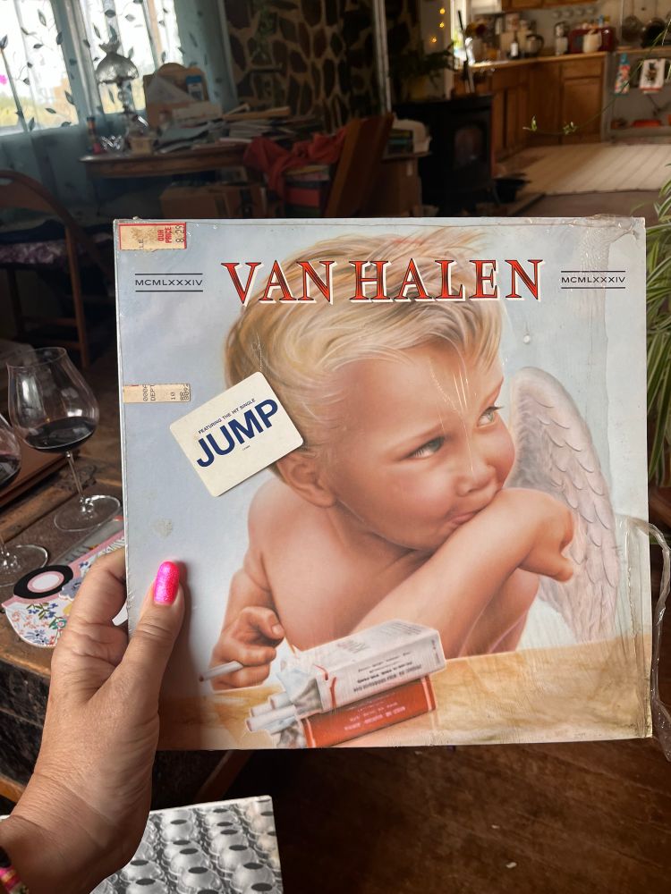 Rachel is holding a copy of the cover of the Van Halen LP which includes Jump and Panama, released in 1984. The cover has a cheryb on the front with wings and a couple of packs of smokes. Rachel’s thumbnail is pink and there is a wineglass on the coffee table in the background behind the album,