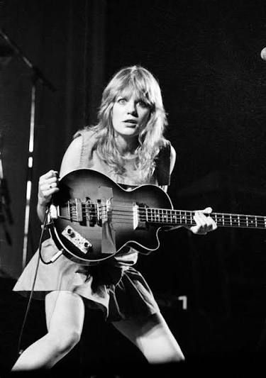 Black and white photo of bassist Tina Weymouth of Talking Heads in the Stop Making Sense concert movie. She has long blonde hair and is wearing a sleeveless mini dress. She’s holding her bass horizontally across her body and is kind of hunched with her knee bent. She has a theatrical expression of surprise or “huh?!”