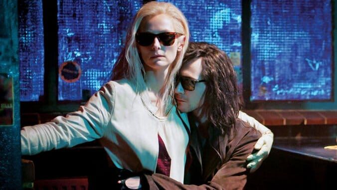 In a scene from Only Lovers Left Alive, Eve (Tilda Swinton) and Adam (Tom Hiddleston) embrace each other in a Detroit bar. Eve is wearing a white jacket and has platinum blonde hair. Adam has long brown hair and is wearing a brown jacket. Both are wearing dark glasses and it’s a dark bar scene. There are windows behind them, with blinds that are open but it’s dark on the other side, though the background light is blue.