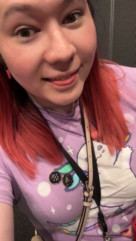 selfie of a woman in an elevator, she has dyed red medium-long hair and is smiling with her tongue poking out slightly. she’s wearing a purple graphic tee with an axolotl drawn on it.