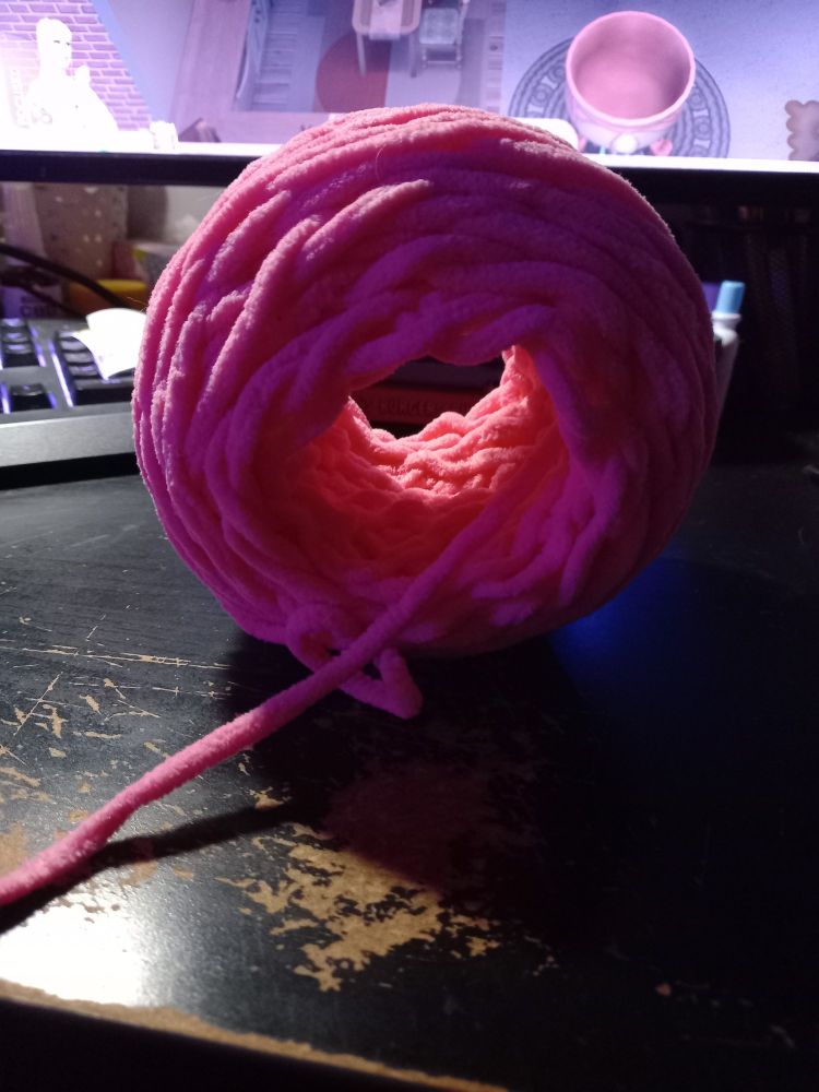 A yarn cake made of chunky pink yarn that's being worked from the middle, sitting on a black computer desk