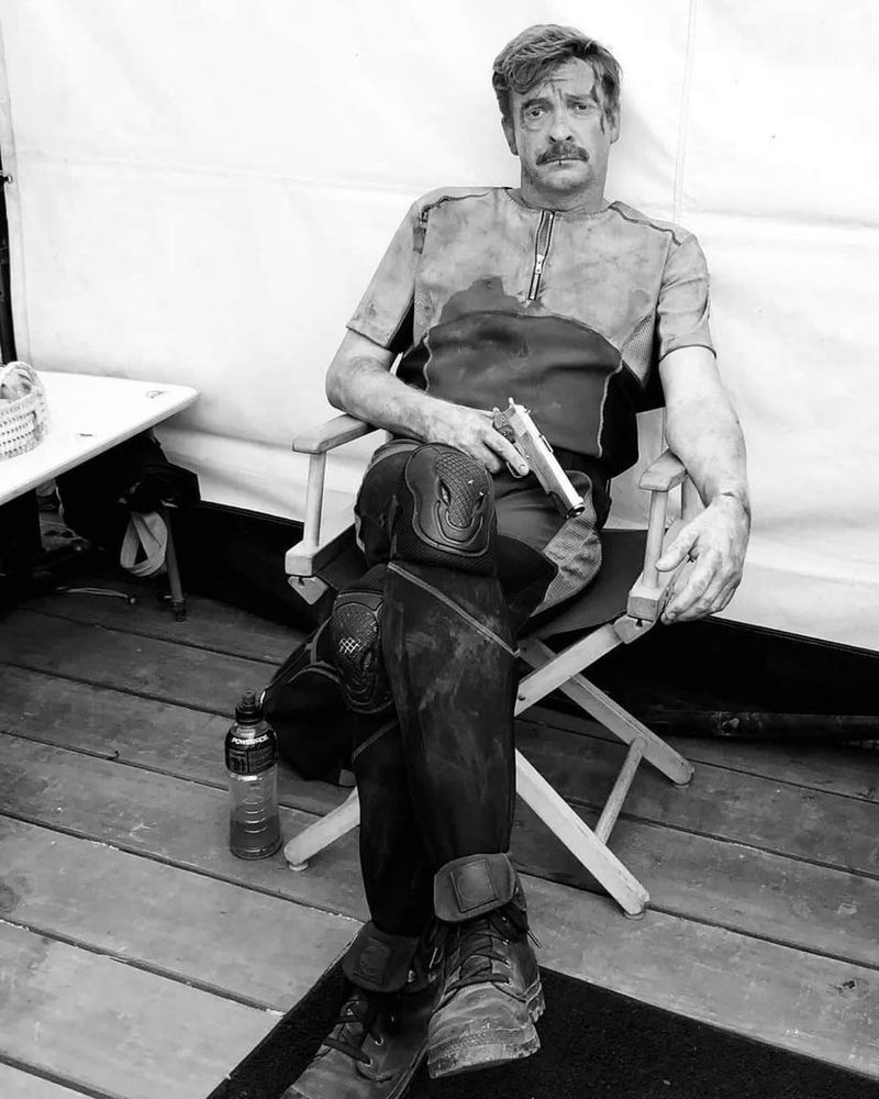 Black and white bts photo of rhys as Steve in wrecked, sitting in a chair with his legs crossed