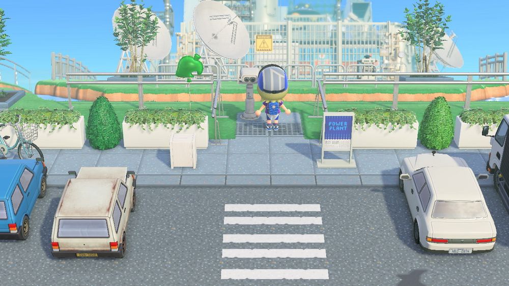 A screenshot from Animal Crossing: New Horizons. An island rep standing in an urban parking lot faces away from the camera, looking at a large power plant structure in the distance.