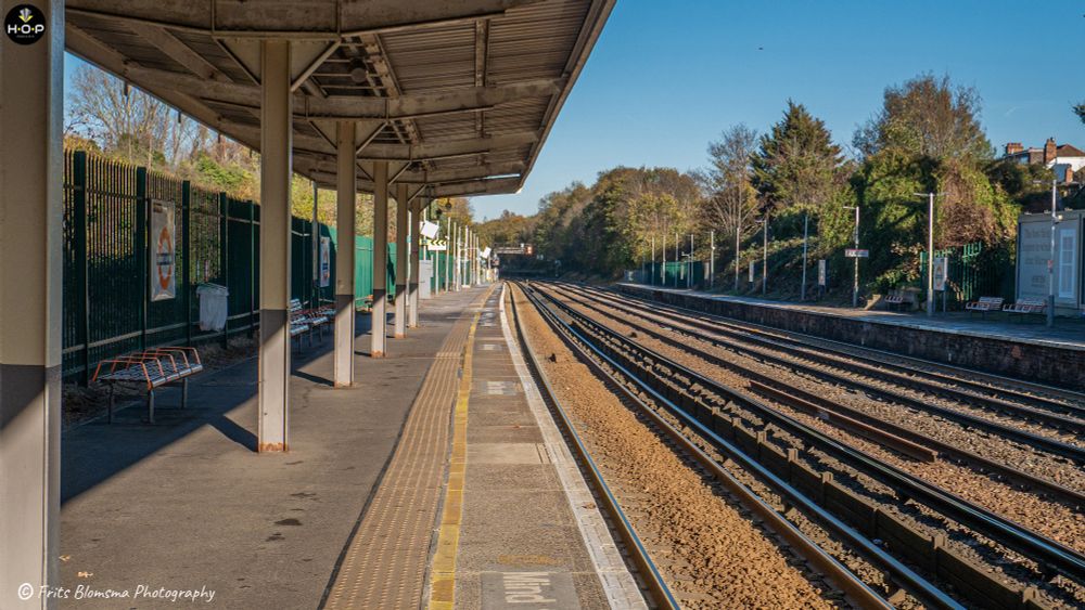 This image captures the view down a typical commuter or suburban railway station in what appears to be the United Kingdom, suggested by the roundel visible on the fence and the style of the station furniture.

The image is well-exposed, with the strong sunlight creating a nice contrast between the shadowed platform and the brightly lit tracks. The lines of the platform and the tracks draw the viewer's eye deeply into the composition, making the railway line a strong leading line in the image.
