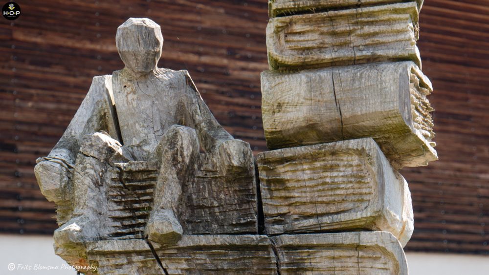 This photo depicts a striking wooden sculpture photographed in Switzerland during the summer. The sculpture appears to be hand-carved from large, stacked wooden beams. It depicts a seated, faceless human figure carved in a minimalist, almost abstract style. The figure is positioned on what seems to be a stack of oversized wooden “books” or blocks, giving the impression of contemplation or reading.

The weathered texture of the wood, with cracks and rough grain, adds a sense of age and organic character to the piece. The background shows a blurred wooden structure, which complements the earthy tones of the sculpture. The lighting suggests it was taken on a bright day, emphasizing the natural textures and shadows.