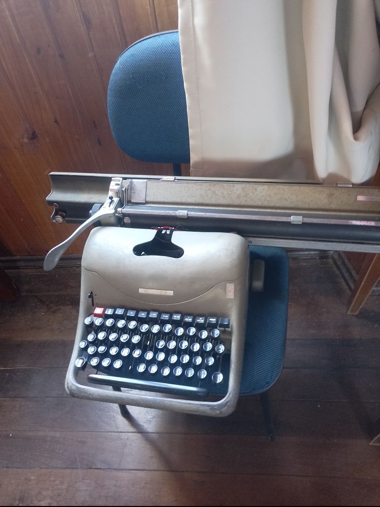 Foto de uma antiga máquina de escrever cinza com botões pretos sobre uma cadeira azul e com um assoalho de madeira