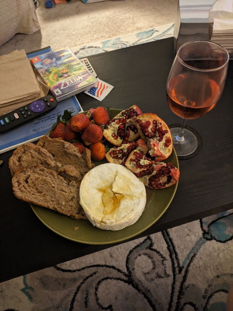 plate of food, containing baked Brie with wildflower honey, gluten free bread, strawberries and a pomegranate. it is accompanied by a glass of red wine