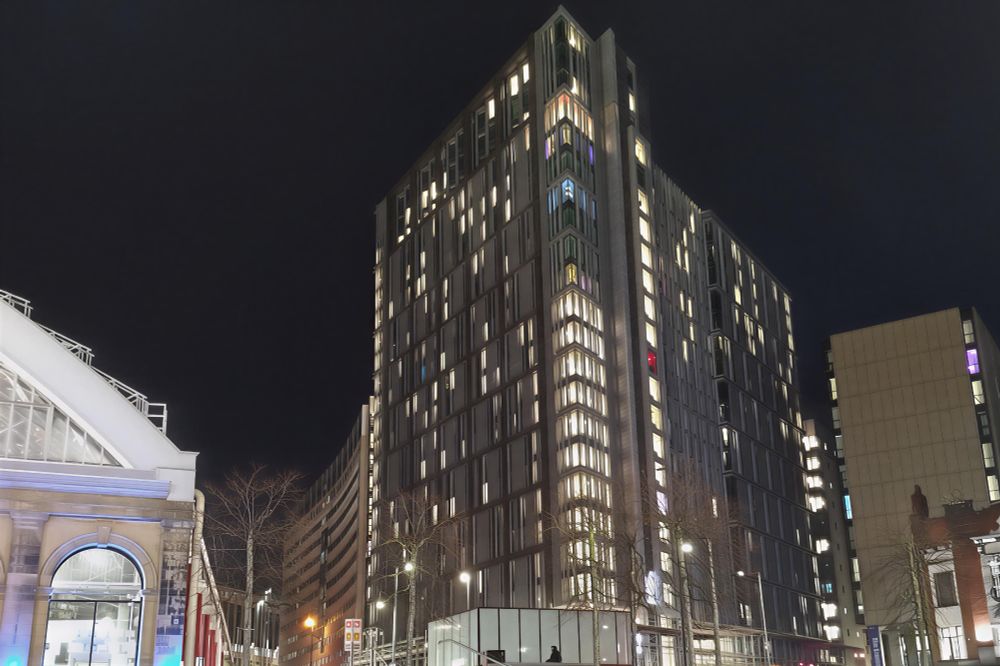 Modern high-rise glowing at night beside a classical building, with bare trees and city lights.