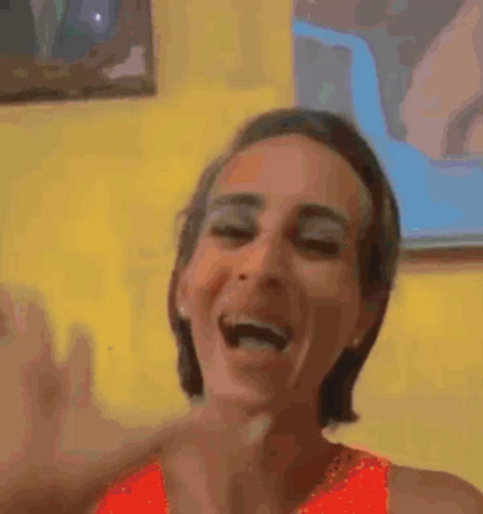 a woman in an orange tank top is laughing and making a funny face in front of a yellow wall .