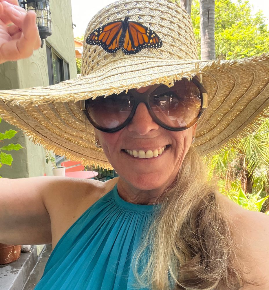 A woman with a monarch butterfly on her hat, she’s wearing a teal blue dress, has a ponytail, garden hat and oversized: tortoiseshell sunglasses 