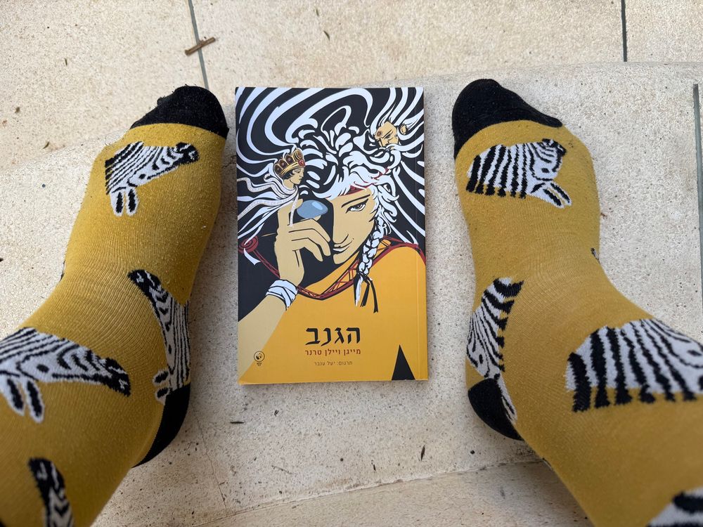 My feet in yellow socks with zebras on them next to the Hebrew edition of “The Thief” by Megan Whalen Turner which has a striking yellow, black and white cover 