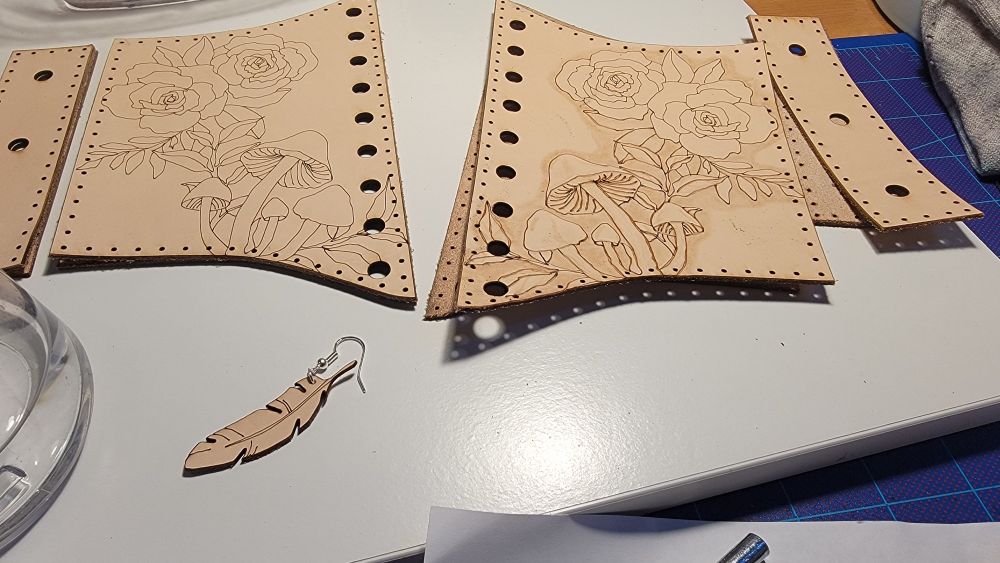 A photo of two incomplete projects. One leather earring and a partially tooled elastic corset design with mushrooms and flowers outlined